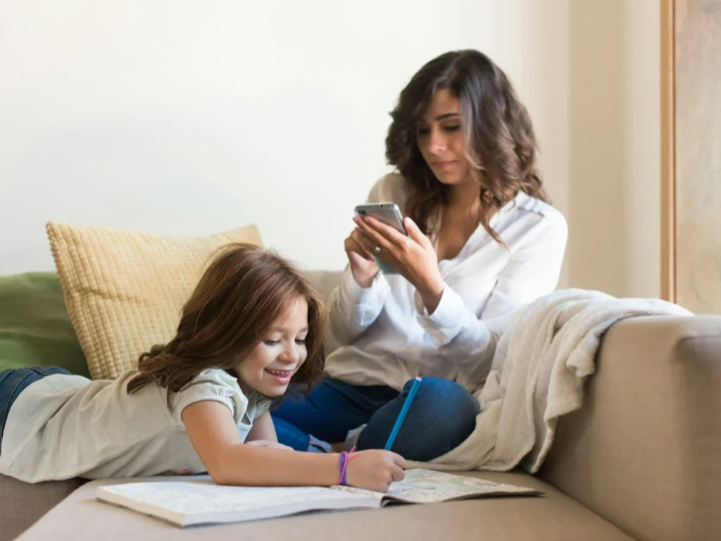 A parent looks down at their smartphone while their child plays alone in the background, illustrating the challenge of being present.