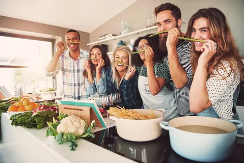 A group of friends laugh together as they help a new parent prepare a meal in the kitchen.