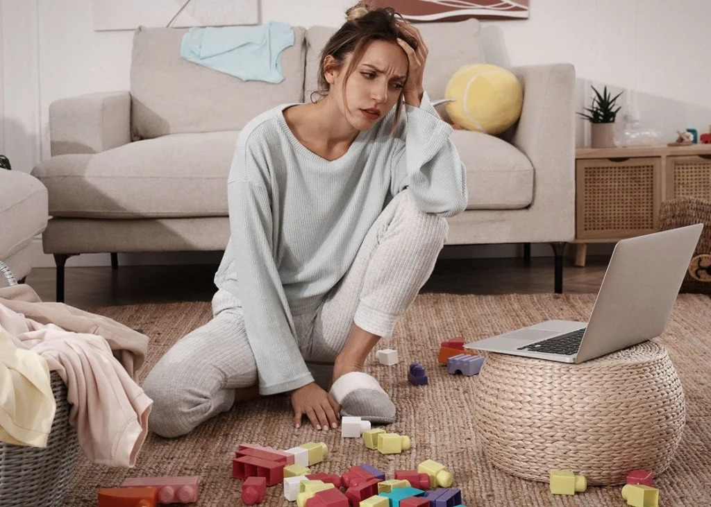A parent sits on the floor surrounded by laundry and toys, looking tired but thoughtful.
