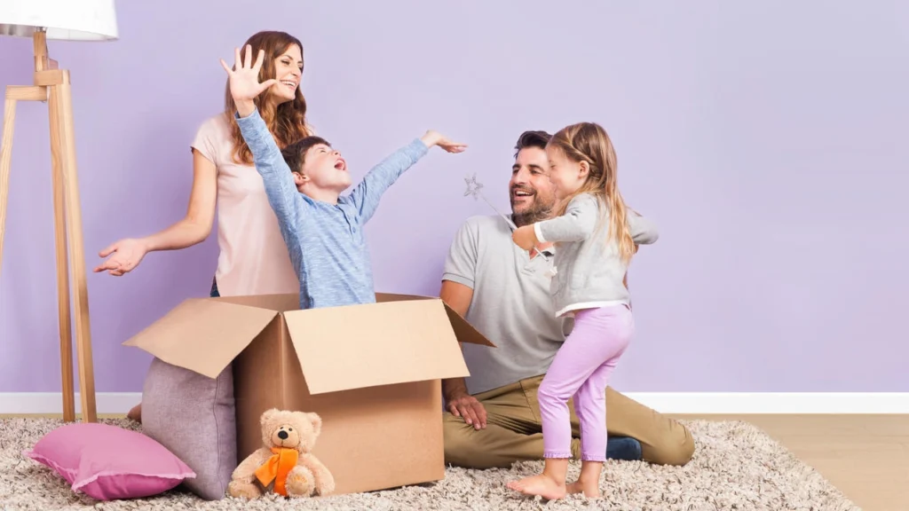 A parent sits at a table and supervises their young child who is playing with clothespins on a cardboard box.
