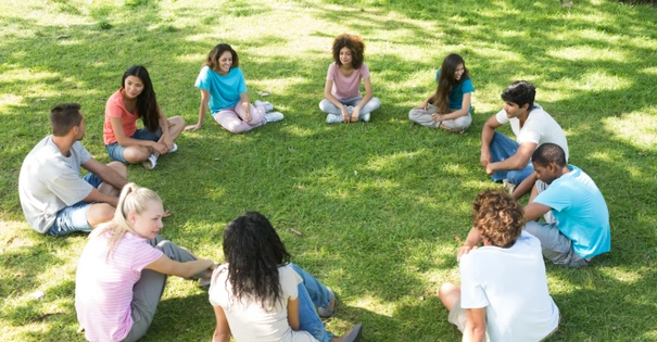  A group of diverse parents and their young children sit in a circle on the grass, building a community.
