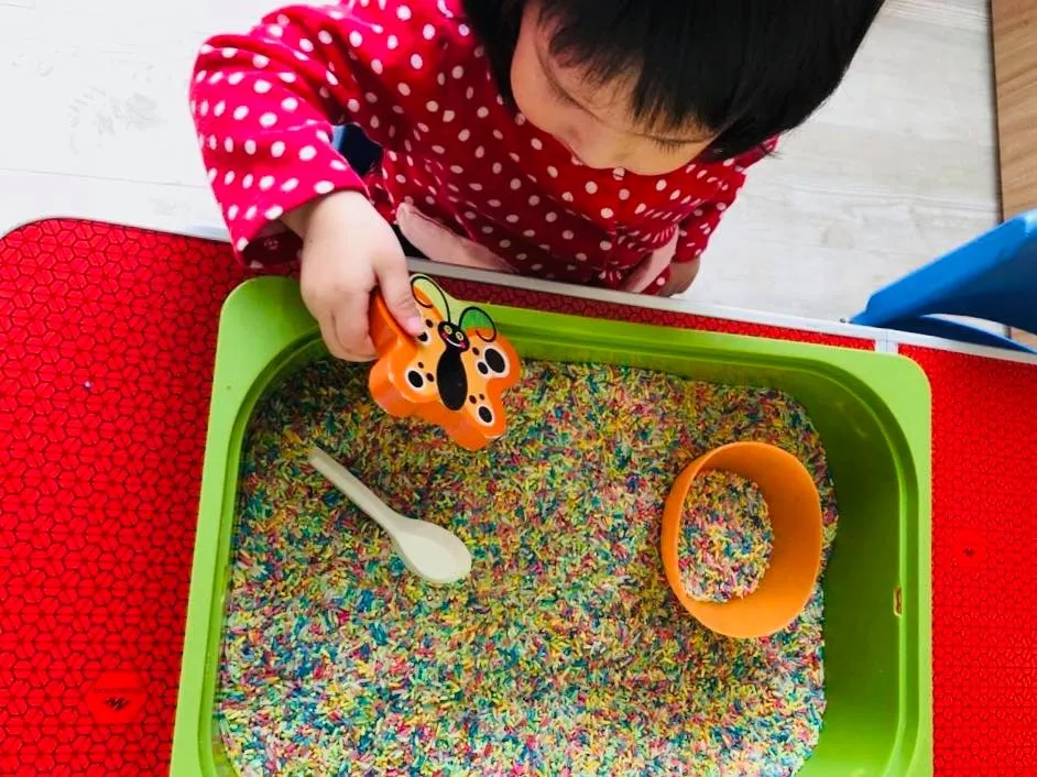 A toddler's hands using a small scoop to move rice into orange bowl for a sensory activity.