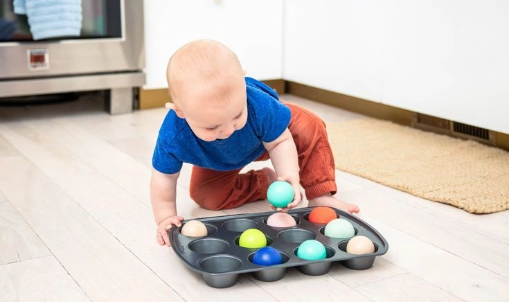 A toddler uses their fingers to sort bright red and blue pom-poms into the cups of a muffin tin.