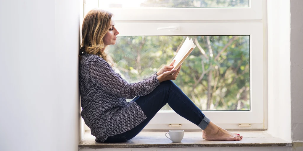 A woman sitting quietly by a window holding a notebook, looking relaxed and calm.