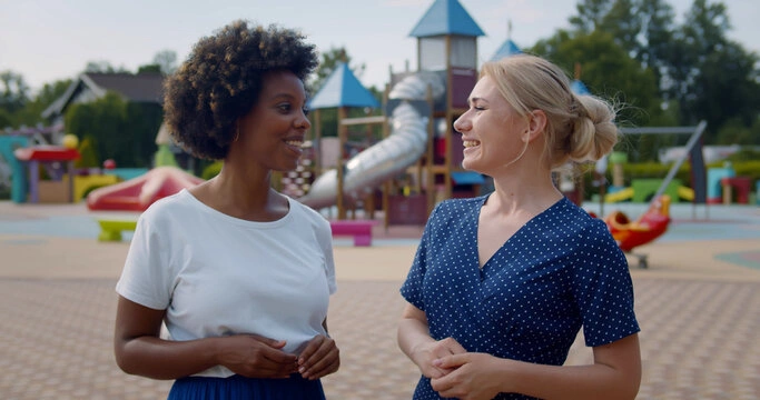 Two mothers greeting each other at a playground while their children play nearby.