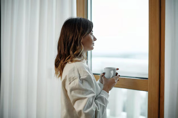 A tired mother holding a cup of tea looking out the window, illustrating parental isolation.