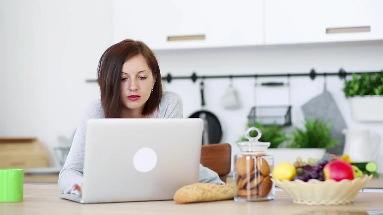 A parent sitting at a kitchen table with a laptop, exploring teletherapy options for parental anxiety, with children's items visible in the background.